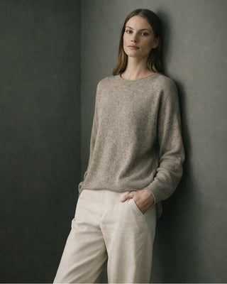 Woman in a taupe sweater and cream trousers leaning against a dark wall