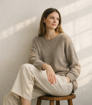 Woman in an oversized taupe sweater and cream trousers seated on a stool