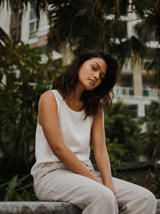Woman in a white sleeveless linen top and beige linen trousers seated outdoors among palms